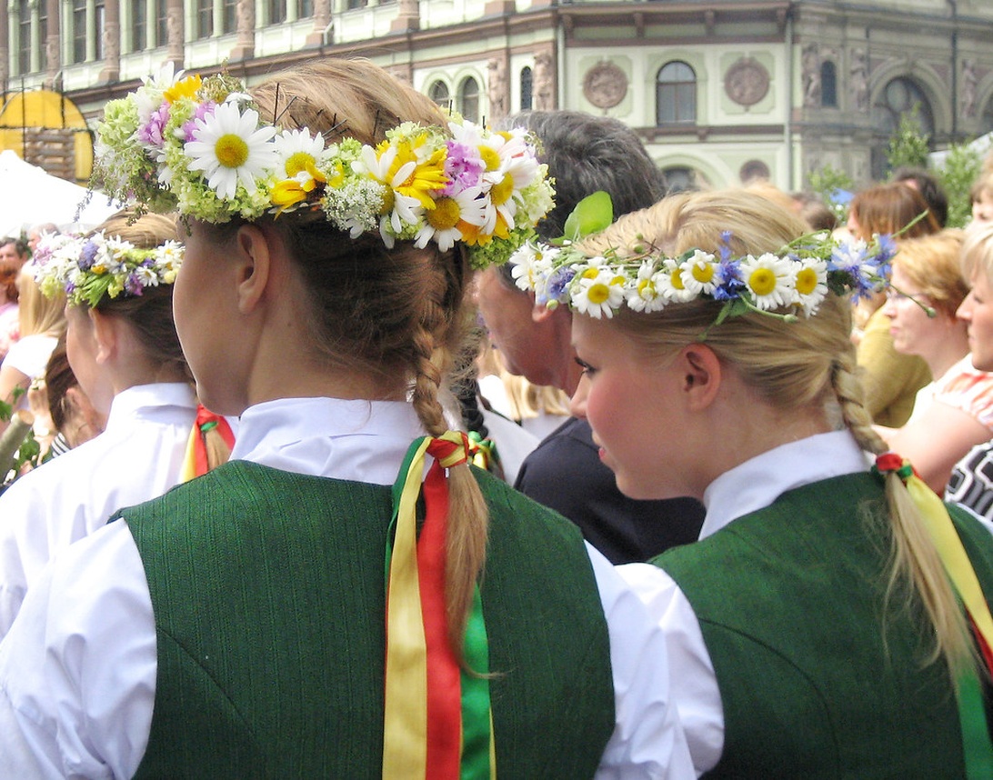 Flera personer bär traditionella blomsterkransar av prästkragar, vildblommor och grönska under ett midsommarfirande. De är klädda i traditionella gröna västar med färgglada band, vilket antyder ett skandinaviskt eller europeiskt midsommarfirande där blomsterkransar är en traditionell prydnad.