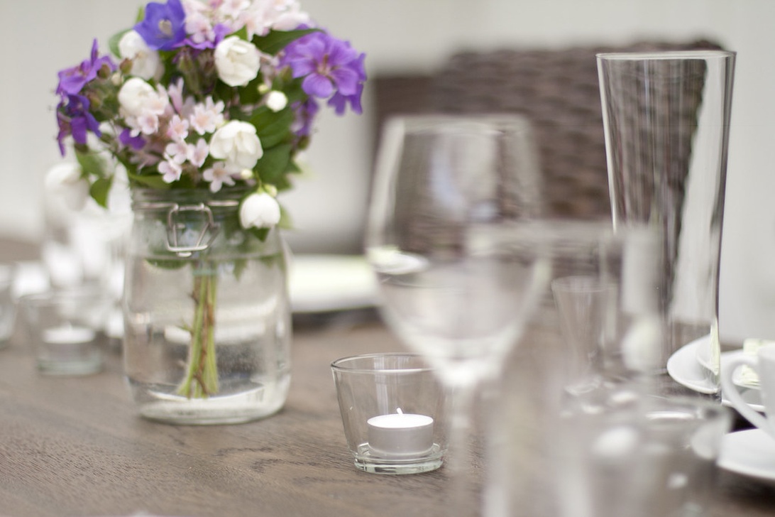 En bordsdukning med en glasburk innehållande en liten bukett vildblommor, inklusive lila blommor, vita blommor och grönska. Bilden visar ett rustikt eller naturligt arrangemang som kan representera blommor som kan användas vid midsommarfiranden.